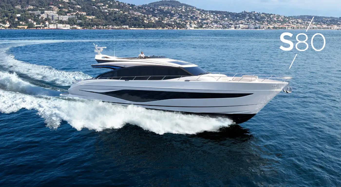 Stylish yacht Princess S80 cruising along a coastal area with a scenic town and hillside in the background.