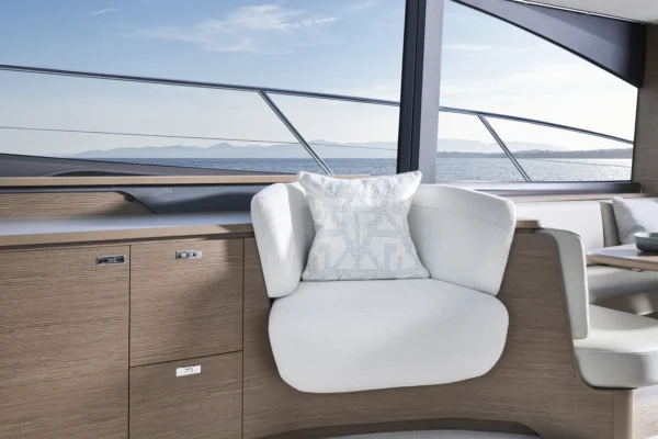 Elegantes Lounge-Bereich-Interieur auf der Princess S65, mit einem komfortablen weißen Sessel, dekorativem Kissen und Holzverkleidung im Satin-Finish. Durch das große Fenster zeigt sich eine Aussicht auf das ruhige Meer und eine ferne Berglandschaft.