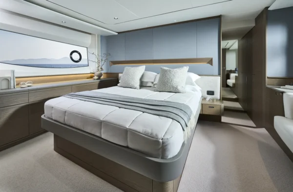 Luxuriöse Owner Kabine auf der Princess S65, ausgestattet mit einem großen, gemütlichen Bett, weichen Kissen und modernen Holzakzenten in Satin. Ein großes Fenster bietet eine beruhigende Aussicht auf das Meer und lässt natürliches Licht in den Raum.