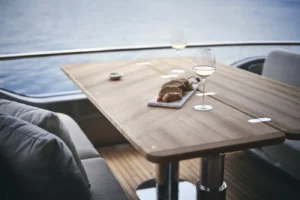 Ein luxuriös gedeckter Esstisch auf einer Princess Y85 Yacht, mit einem Holztisch, auf dem ein Teller mit Brot und zwei Gläser Weißwein stehen, mit Blick auf ruhige, blaue Gewässer